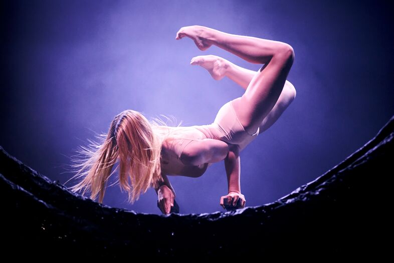 Een naakte vrouw voert een bruisende acrobatische pose uit op een futuristische achtergrond met paarse tinten.
