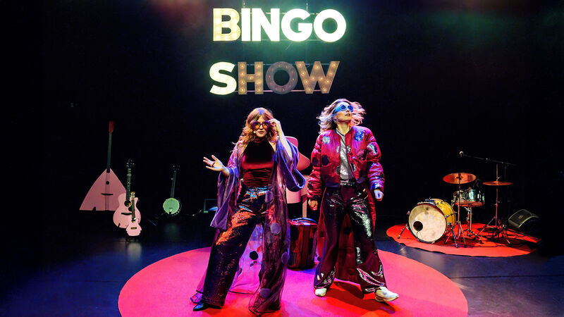 Twee performers op een podium met een 'Bingo Show'-decor en muzikale instrumenten op de achtergrond.