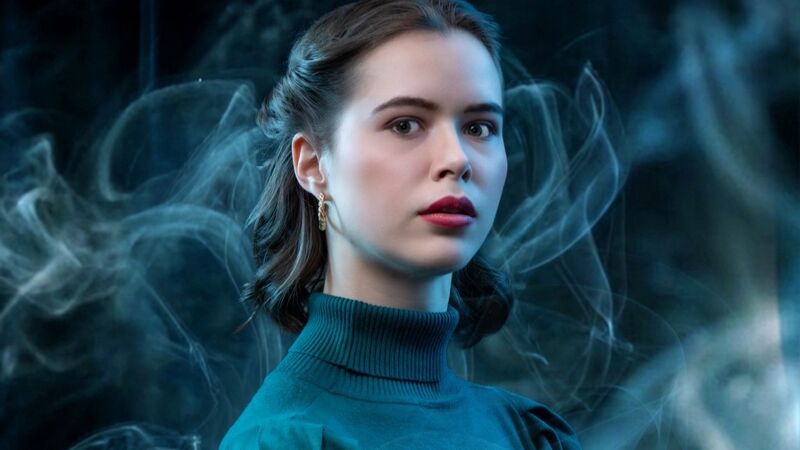 Een vrouw met een lichte huid en lang bruin haar, gekleed in een groene trui, staat in een mysterieuze, nevelachtige omgeving.