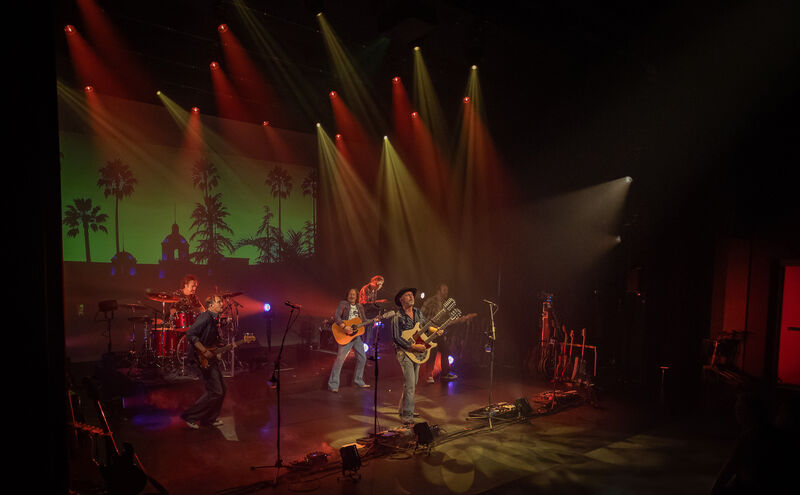 Een band speelt live op een podium met kleurrijke verlichting en een achtergrond van silhouetten van bomen.