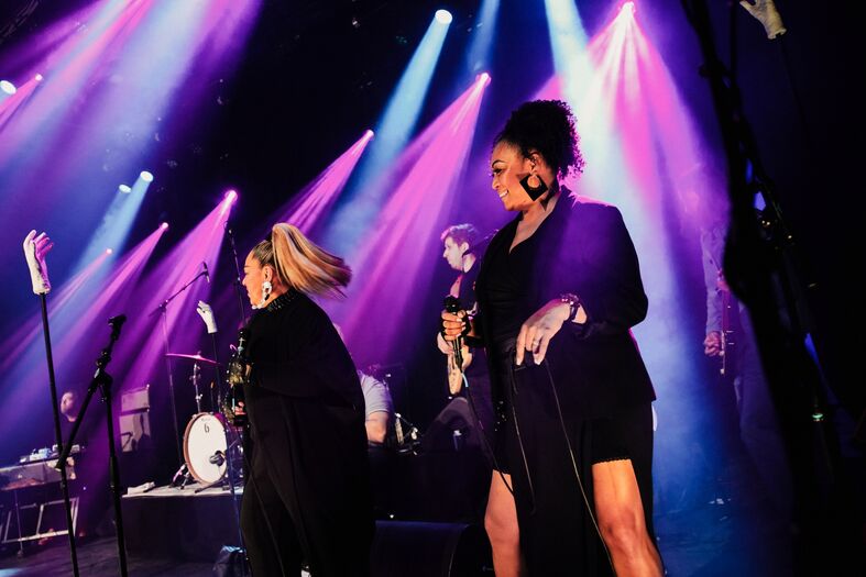 Vrouwen zingen op een podium met kleurrijke lichten, omringd door muzikanten in een live muziekevenement.