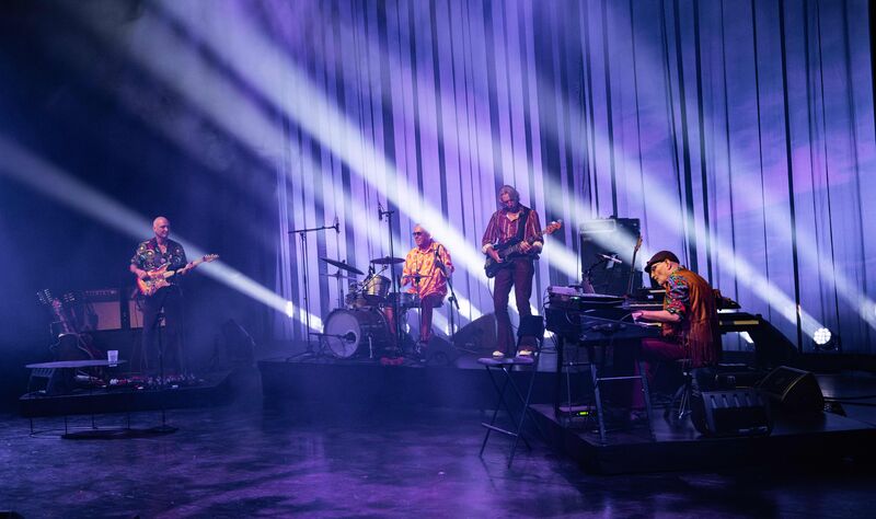 Muzikanten optreden op een podium met paarse lichten en een theatrale achtergrond.