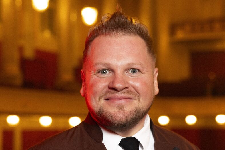 Alexander Wurz met een vrolijke uitdrukking, gekleed in een donker jasje en stropdas, met een concertzaalachtergrond.