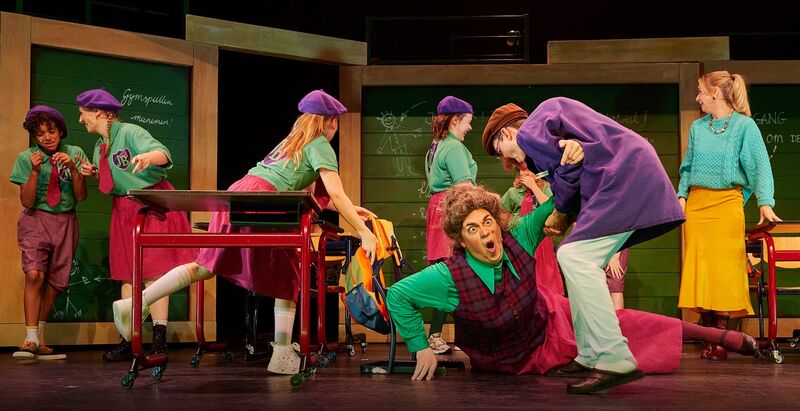 Theateruitvoering met zeven acteurs in kleurrijke kostuums, in een humoristische scène met interactie en beweging.