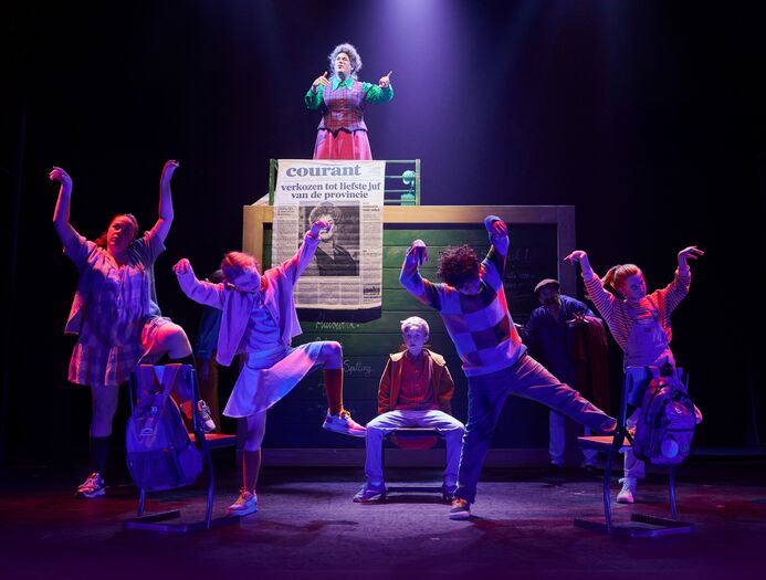 Theaterproductie met acteurs in kleurrijke kostuums, een centrale figuur op een verhoging en anderen in beweging.