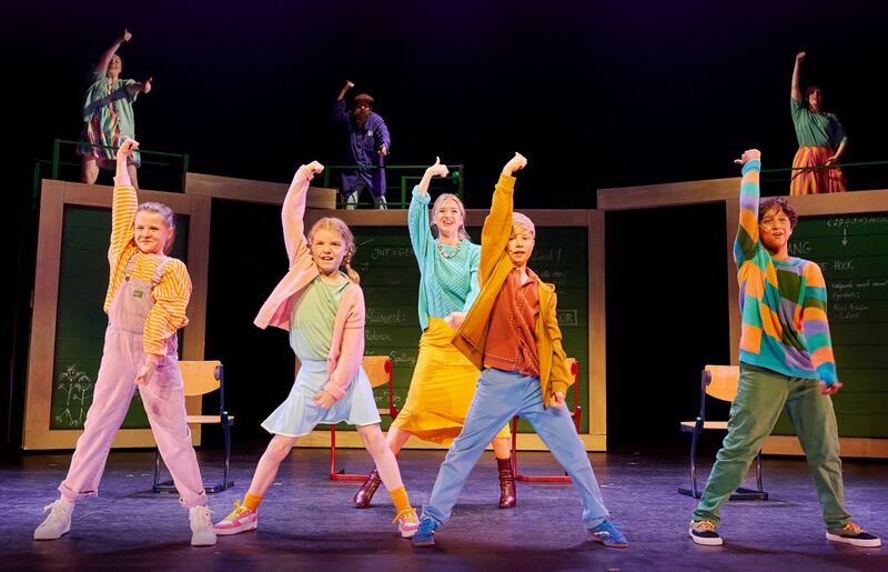 Groep dansers op het podium in kleurrijke kleding, met een energieke houding en een vrolijke choreografie.