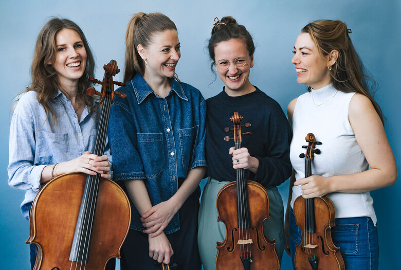 Groep van vier vrouwen met strijkinstrumenten, staand en lachend voor een blauwe achtergrond.