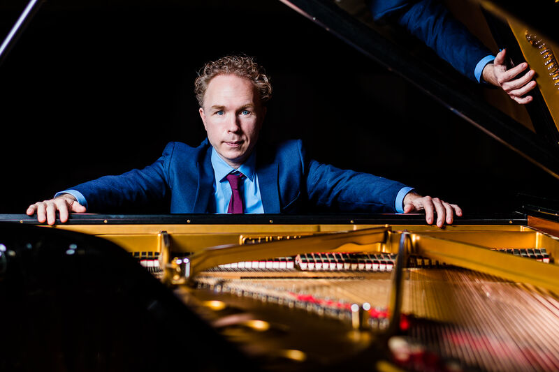Man in een blauw pak zit aan een open vleugel, met handen op de rand van de piano, kijkend naar de toetsen.
