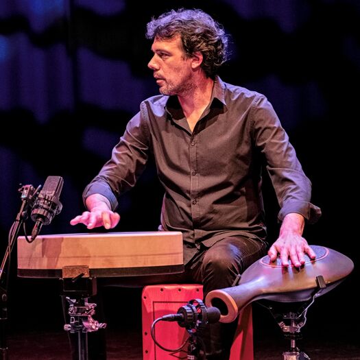 Mannetje speelt muziekinstrumenten, zittend op een podium met microfoons en een artistieke achtergrond.