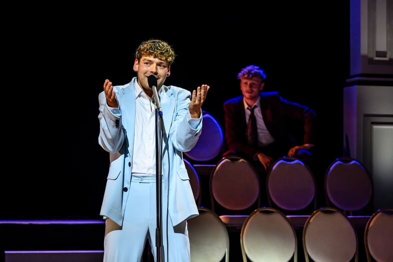 Een jonge man in een lichtblauw kostuum zingt op een podium, terwijl een andere man op de achtergrond zit.
