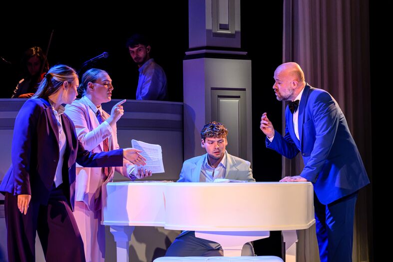 Vijf acteurs op een podium, met een piano, in een dramatische scène van een theaterstuk.