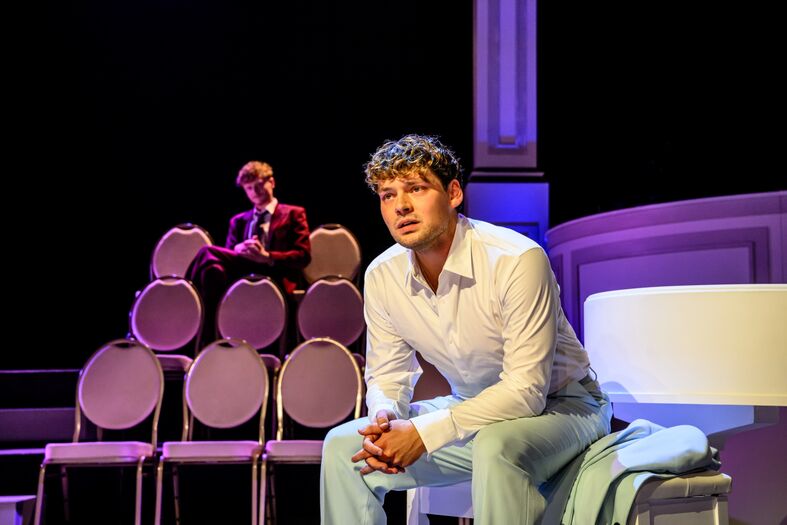 Een acteur in een wit overhemd zit op een stoel, met een andere acteur op een stapel stoelen op de achtergrond.