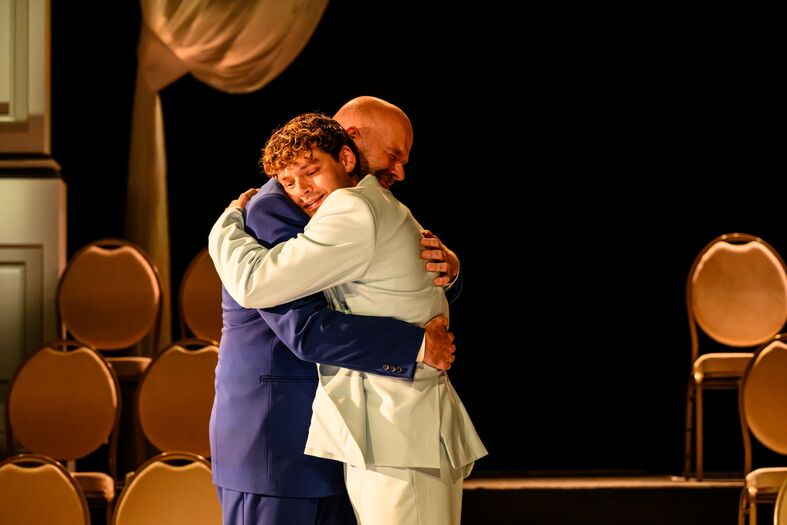 Twee mannen omarmen elkaar emotioneel op een podium, met stoelen op de achtergrond.
