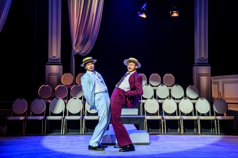 Twee dansers in formele kleding voeren een choreografie uit op een voorstelling met lege stoelen op de achtergrond.
