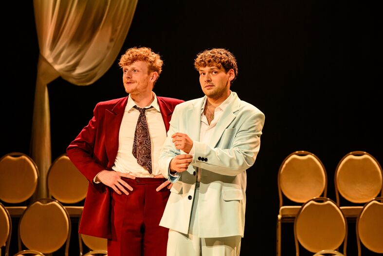 Twee mannen op een podium, gekleed in opvallende outfits, staan naast elkaar voor een rij stoelen.