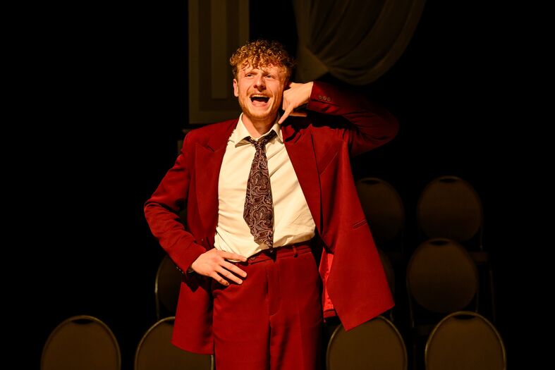 Een man in een rood pak staat op een podium en zingt, terwijl hij naar het publiek wijst.