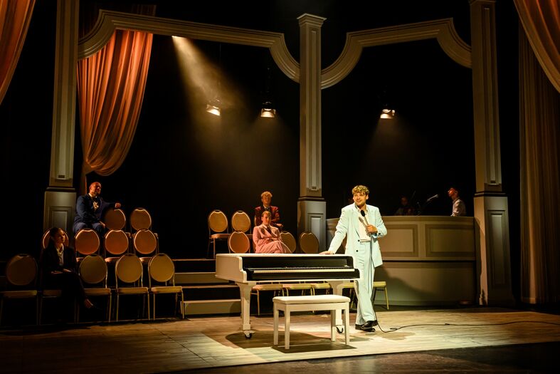 Man in lichtgekleurde kostuum zingt achter een piano op een theatrale scène met stoelen en verlichting.