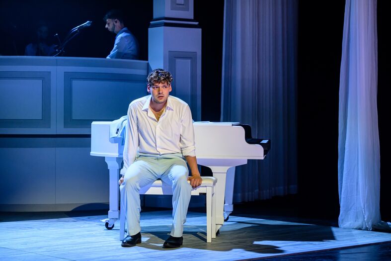 Een man in een wit overhemd zit op een pianokruk, naast een witte piano, met een melancholische uitstraling.