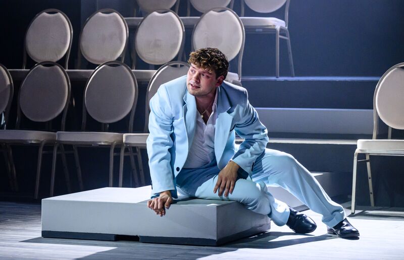 Een man in een lichtblauw kostuum zit op een podium met stoelen op de achtergrond, met een expressieve houding.