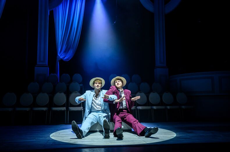 Twee acteurs in kostuums zitten op een podium met een spotlight, omringd door stoelen in de achtergrond.