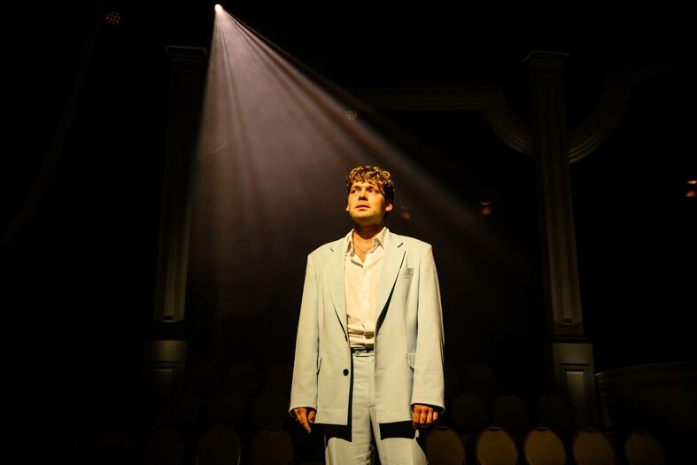 Een jonge man in een lichtblauwe outfit staat onder een enkele lichtstraal op een podium, met een onduidelijke achtergrond.