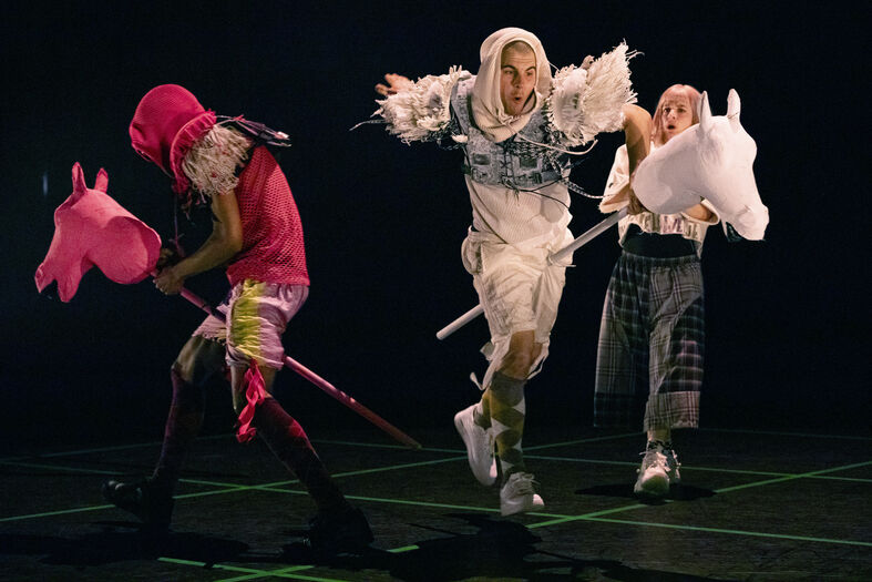 Drie performers in kleurrijke outfits, met fantasierijke accessoires, voeren een dynamische dansroutine op een donkere achtergrond uit.