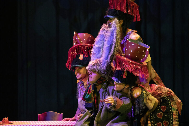 Drie performers in kleurrijke kostuums met grote hoeden en een opvallende baard voeren een theatrale act op.