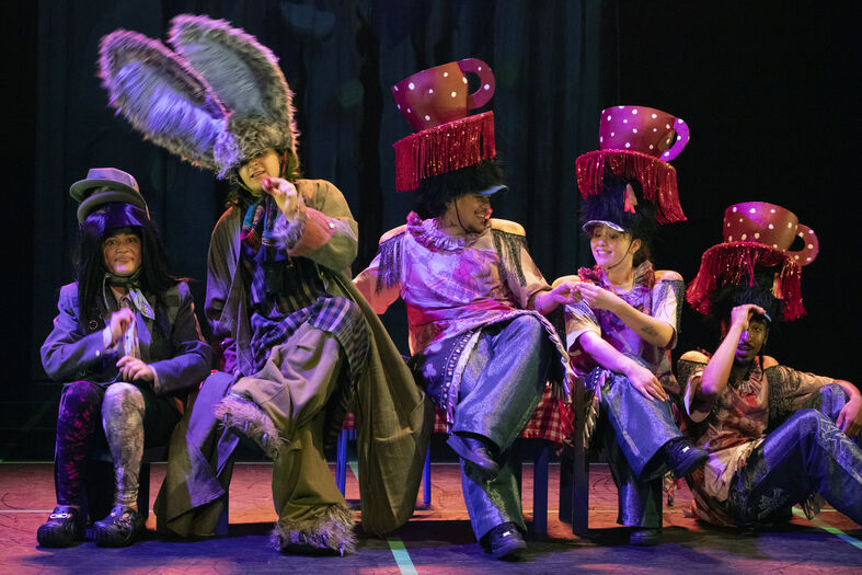 Vijf acteurs in kleurrijke kostuums met hoeden, die op een podium een theatrale scène spelen.