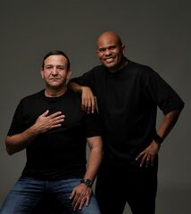 Najib Amhali zit terwijl Roué Verveer achter hem staat, beiden in zwart voor grijze achtergrond.