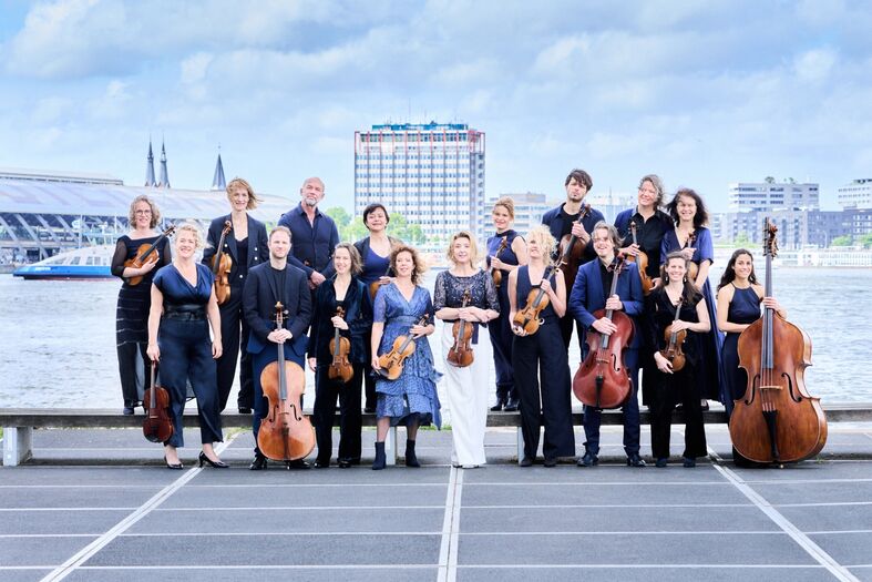 Groep muzikanten in formele kleding poseert op een pier met uitzicht op een stad, met verschillende instrumenten in handen.