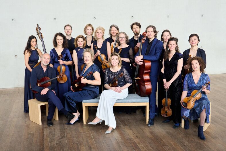 Groepsportret van een symfonieorkest, bestaande uit 20 musici in formele kleding, zittend en staand in een moderne ruimte.