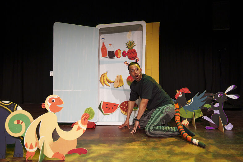 Kindervoorstelling met acteur bij een koelkast vol kleurrijke papieren fruit, omringd door cartoonachtige dieren.