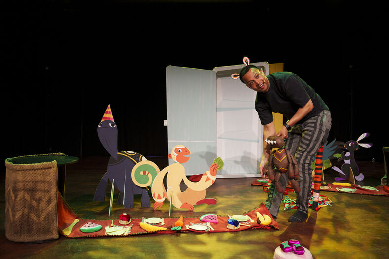 Een performer staat op een podium naast kleurrijke, papieren dieren en decorstukken, met een vrolijke, speelse sfeer.