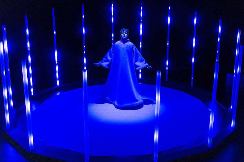Een figuur in een blauwe outfit staat centraal op een ronde constructie, omgeven door blauwe verlichting.