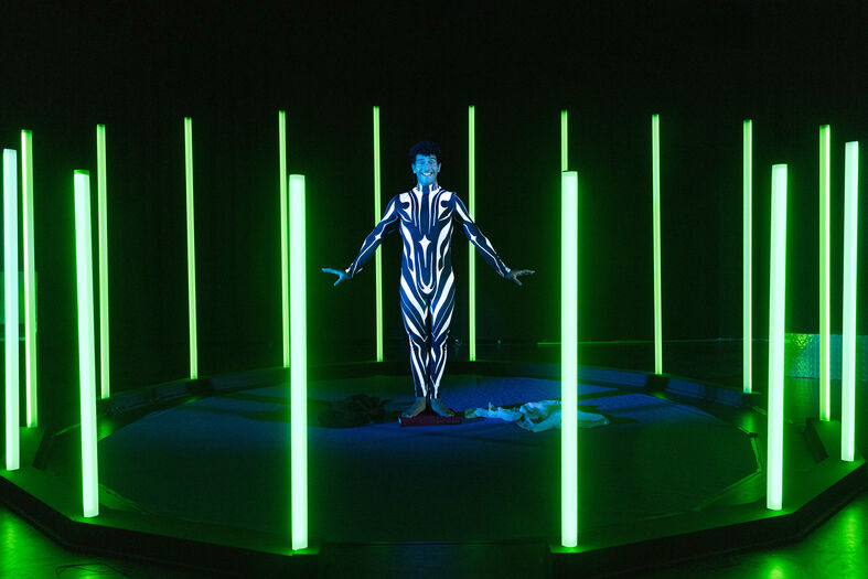 Een menselijk figuur in een futuristische outfit staat omringd door groene, lichtgevende staven in een donkere ruimte.