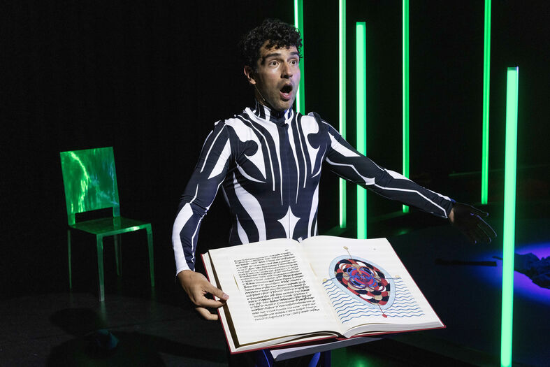 Man in een zwart-wit kostuum staat met een groot geopend boek, omringd door groene lichtstralen.