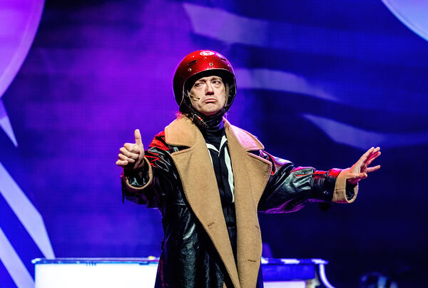 Performende artiest in een leren jas en helm, met uitgestrekte armen, op een kleurrijk verlichte achtergrond.