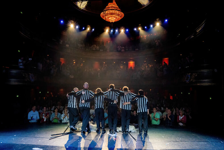 Zes scheidsrechters staan op een podium met fel licht, terwijl het publiek in een theater op hen kijkt.