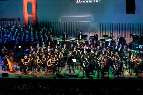 Een orkest speelt op een podium, met muzikanten in zwarte kleding, onder kleurrijke lichten en een groot scherm op de achtergrond.