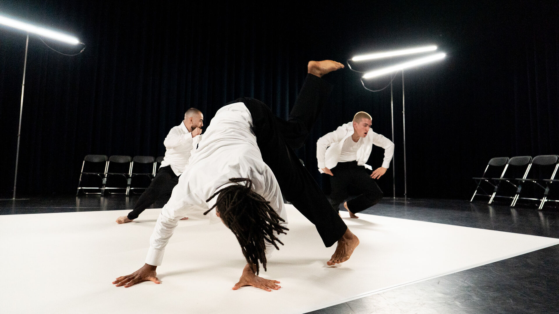 Drie dansers op een podium, één danser voert een acrobatische beweging uit terwijl de anderen toekijken.