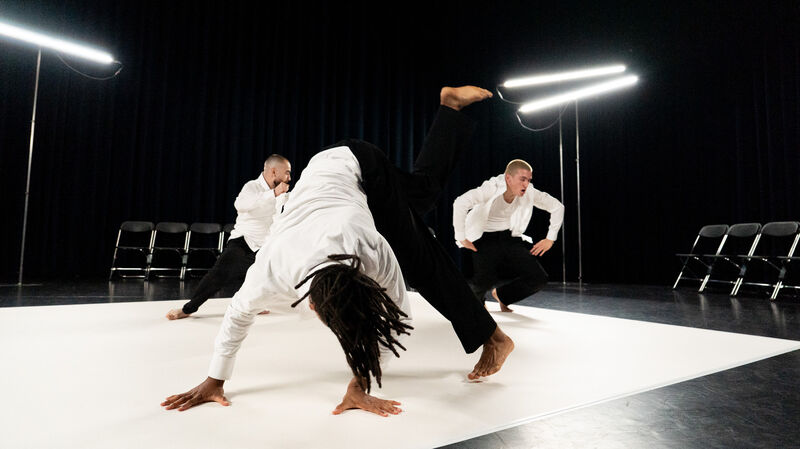 Drie dansers op een podium, één danser voert een acrobatische beweging uit terwijl de anderen toekijken.