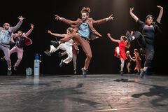 Grote groep dansers springt gelijktijdig omhoog op een podium, met benen en armen dynamisch de lucht in gericht.