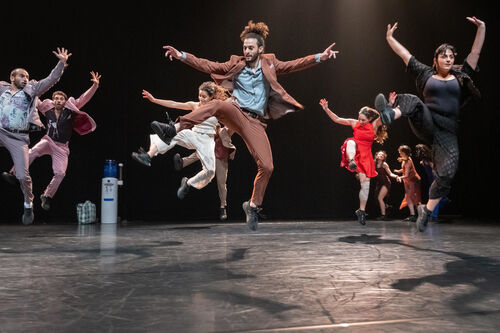 Grote groep dansers springt gelijktijdig omhoog op een podium, met benen en armen dynamisch de lucht in gericht.