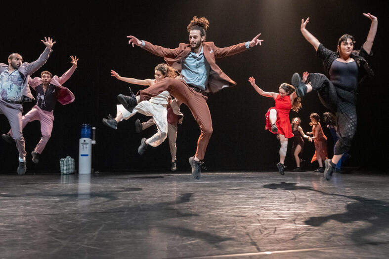 Grote groep dansers springt gelijktijdig omhoog op een podium, met benen en armen dynamisch de lucht in gericht.