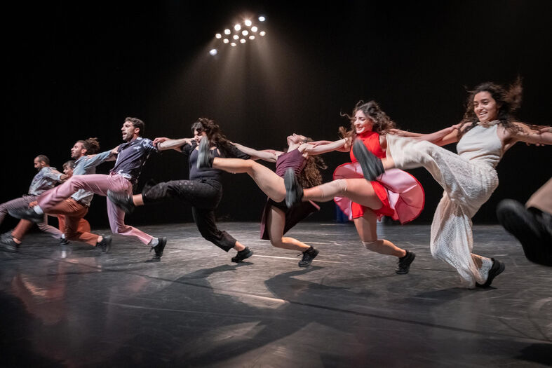 Rij dansers houdt elkaars handen vast terwijl zij synchroon zijwaarts trappen op een verlicht podium.
