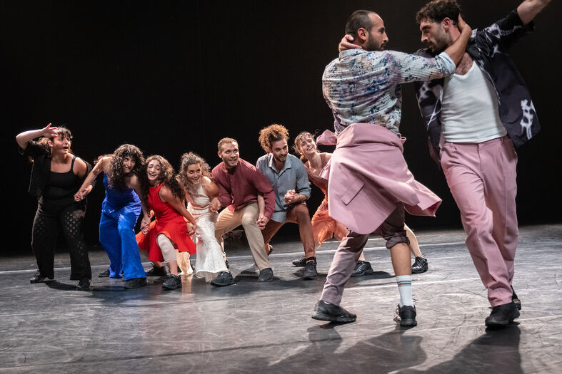 Groep dansers gehurkt in een rij op het podium terwijl twee performers ervoor in een draaiende beweging elkaars armen vasthouden