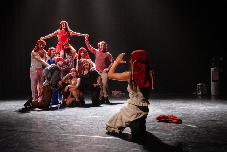 Groep dansers vormt een menselijke piramide op een podium, terwijl een performer vooraan een gebaar maakt richting de groep.