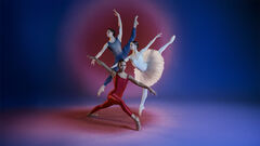Drie balletdansers in rode, witte en blauwe outfits poseren in uitgestrekte balansas tegen een roze‑blauwe achtergrond.