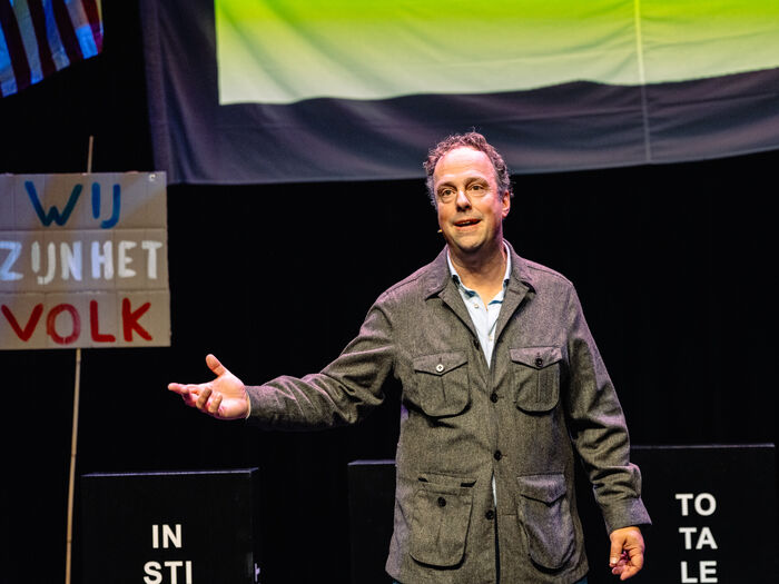 Man op het podium met een microfoon, naast een bord met de tekst "Wij zijn het volk", tijdens een voorstelling.