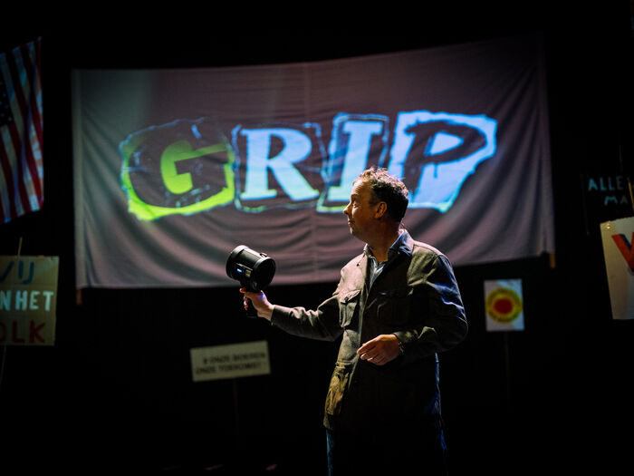 Man op een podium houdt een camera vast voor een groot scherm met de tekst "GRIP" in graffiti-stijl.